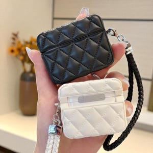 Puede incluir: Se muestran dos fundas acolchadas para AirPod, una negra y otra blanca. Las fundas tienen un patrón de diamantes y una correa para la muñeca. La funda negra está abierta, revelando el interior. La funda blanca está cerrada.