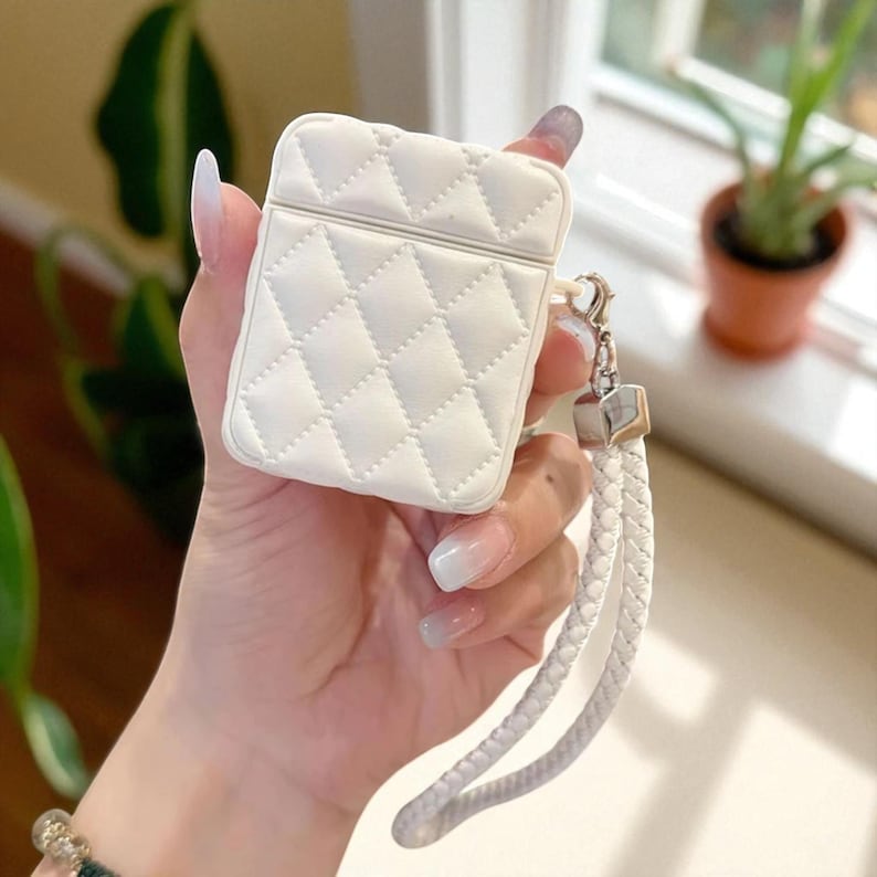 May include: A white protective case for wireless earbuds featuring a quilted diamond pattern on the exterior. The case includes a silver-toned metal clasp attached to a white braided wrist strap, providing a secure way to carry the device.