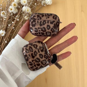 Peut inclure: Deux étuis pour AirPods avec un imprimé léopard. Les étuis sont marron avec des taches marron foncé. L'un des étuis est doté d'un mousqueton argenté avec un charm en forme de cœur noir et une petite étiquette. Les étuis sont sur une surface marron clair.