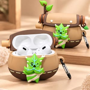 Puede incluir: Una funda de silicona marr&oacute;n y beige para auriculares con un personaje verde en forma de hoja en la parte frontal. El estuche tiene una tapa abatible, un mosquet&oacute;n de metal en el lateral y una peque&ntilde;a rama decorativa con hojas arriba.