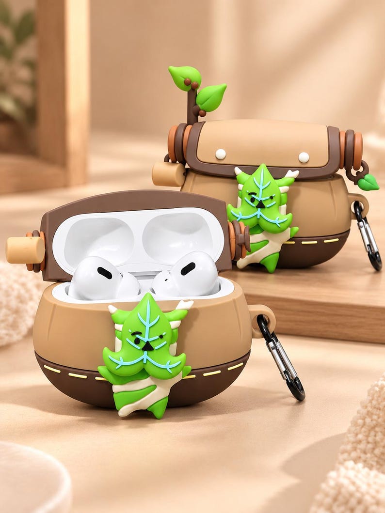 Puede incluir: Una funda de silicona marr&oacute;n y beige para auriculares con un personaje verde en forma de hoja en la parte frontal. El estuche tiene una tapa abatible, un mosquet&oacute;n de metal en el lateral y una peque&ntilde;a rama decorativa con hojas arriba.