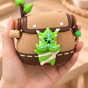 Puede incluir: Una funda protectora de silicona marr&oacute;n para auriculares inal&aacute;mbricos con una solapa marr&oacute;n oscuro, una peque&ntilde;a ramita de hojas verdes en la parte superior izquierda y un dise&ntilde;o central en 3D de una criatura verde con forma de hoja.