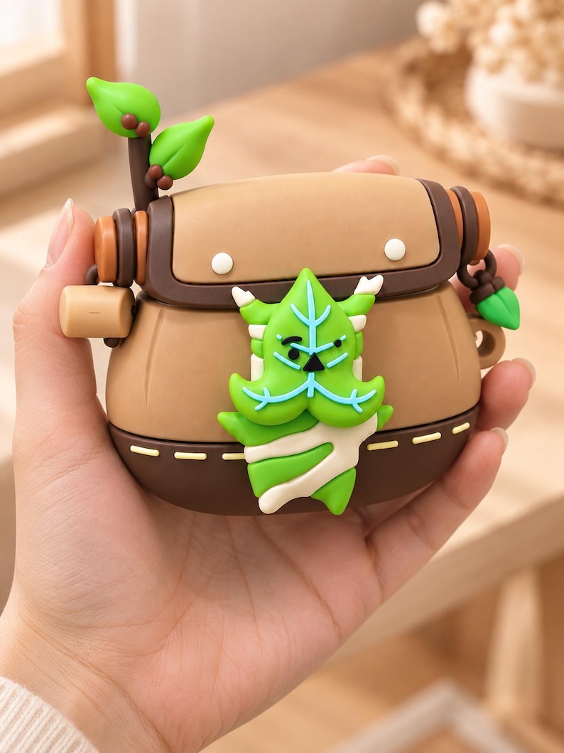 Puede incluir: Una funda protectora de silicona marr&oacute;n para auriculares inal&aacute;mbricos con una solapa marr&oacute;n oscuro, una peque&ntilde;a ramita de hojas verdes en la parte superior izquierda y un dise&ntilde;o central en 3D de una criatura verde con forma de hoja.