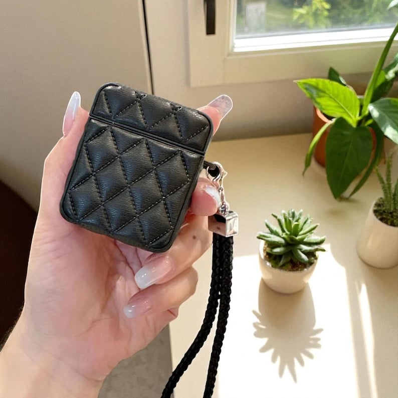 May include: A black quilted protective case for wireless earbuds featuring a diamond-stitched pattern. The case includes a silver-toned metal attachment point connected to a long, thin, black braided wrist strap for carrying convenience.