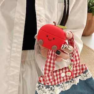 Puede incluir: Una funda roja con forma de manzana para auriculares inalámbricos con una cara sonriente, mejillas marrones y una hoja verde. La funda está sujeta a una correa a cuadros rojos y blancos con una bolsa de mezclilla y encaje.