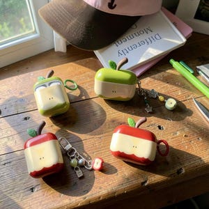 Può includere: Quattro custodie protettive in silicone per auricolari wireless, progettate come mele tagliate. Due custodie sono verdi e due rosse, ognuna con un centro color crema e piccoli semi marroni. Alcune includono portachiavi in metallo attaccati.