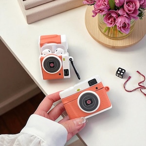 Könnte beinhalten: Zwei orangefarbene und weiße AirPod-Hüllen in Kameraform. Eine Hülle ist geöffnet und zeigt weiße Ohrhörer. Die andere Hülle wird von einer Hand gehalten. Die Hüllen haben ein schwarzes Objektiv und einen kleinen schwarzen Riemen. Ein kleiner schwarzer Würfel und eine Vase mit rosa Blumen sind im Hintergrund.