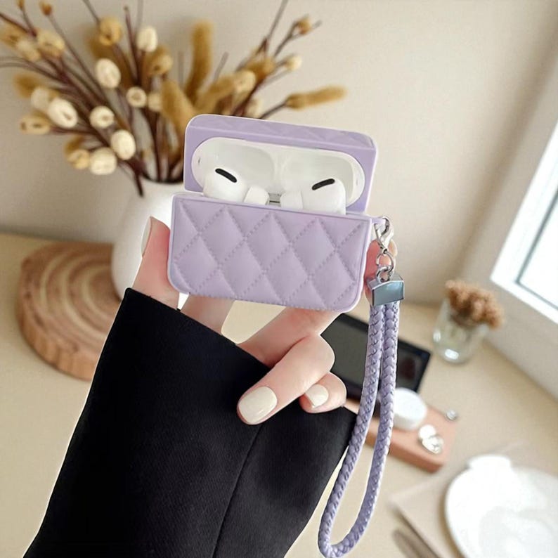 May include: A light purple, quilted silicone case for wireless earbuds, featuring a hinged lid and a matching braided wrist strap attached with a silver-toned metal clasp. The case is open, revealing white wireless earbuds inside the charging unit.
