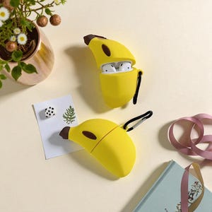 Puede incluir: Dos fundas amarillas con forma de plátano para auriculares inalámbricos. Una funda está abierta, revelando auriculares blancos en su interior. Las fundas tienen puntas marrones y correas negras. El fondo incluye una pequeña planta en una maceta dorada, una tarjeta pequeña y un libro.