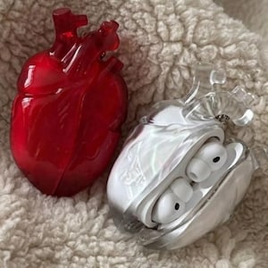 Puede incluir: Dos estuches en forma de corazón. Uno es rojo translúcido, el otro es transparente y contiene auriculares blancos. Los estuches están diseñados para parecerse a corazones anatómicos. Los auriculares son blancos.