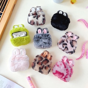 Puede incluir: Una colección de fundas AirPod esponjosas en varios colores y patrones. Las fundas tienen forma de animales, incluyendo conejos y una rana, con pelo suave y texturizado. Los colores incluyen negro, rosa, gris y estampado de leopardo.