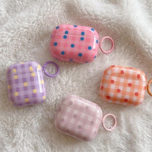 May include: Four colorful AirPod cases with a gingham pattern and polka dots. The cases are pink, purple, orange, and light pink. Each case has a matching ring for attaching to a keychain or bag.