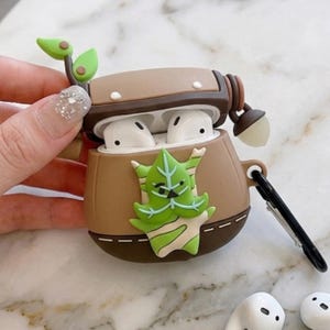 May include: A brown and tan protective case for wireless earbuds, with a green leaf-like character on the front. The case has a hinged lid and a black carabiner clip. The earbuds are white.