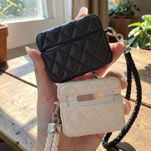 May include: A black and a white protective case for wireless earbuds, both featuring a quilted diamond pattern. The black case includes a black braided wrist strap, while the white case features a silver-toned metal hinge and a white braided wrist strap.