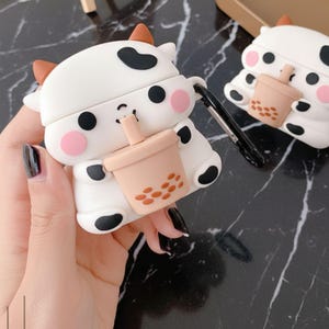 May include: White and black cow-shaped AirPod case with brown horns and black spots. The case has pink cheeks and is holding a peach-colored bubble tea. A black carabiner clip is attached to the case.