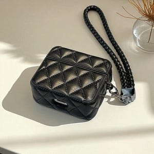 May include: A black protective case for wireless earbuds featuring a quilted diamond pattern with visible stitching. The case includes a matching black braided wrist strap attached to a silver-toned metal clasp on the side of the rectangular housing.