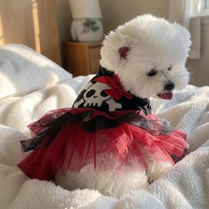 Peut inclure: Un petit chien blanc au pelage duveteux portant un gilet noir orné d'un motif de crâne et d'os croisés blancs. Le gilet présente un nœud rouge à l'encolure et une jupe superposée en tulle rouge et noir. Le chien a des yeux sombres et la langue rose.