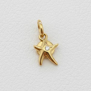 Puede incluir: Colgante de estrella de mar dorada con un pequeño diamante en el centro. El colgante tiene un lazo para una cadena. El diseño de la estrella de mar es detallado y pulido. Fondo blanco texturizado.
