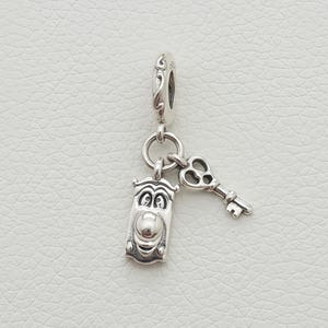 Könnte beinhalten: Ein silberner Charm-Anhänger mit einem rechteckigen Gesicht und einem Schlüssel. Der Charm-Anhänger ist an einer dekorativen Schlaufe befestigt. Der Schlüssel hat ein detailliertes Design. Der Charm ist wahrscheinlich für ein Armband oder eine Halskette.