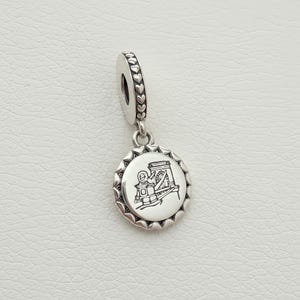 Puede incluir: Un charm de plata con una ilustración detallada de un puente en un colgante redondo. El colgante tiene un borde decorativo y cuelga de una fianza con recortes en forma de corazón. El charm está diseñado para una pulsera o collar.