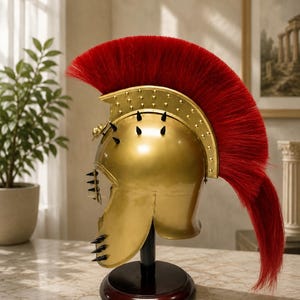 May include: A gold-colored metal helmet featuring a prominent, vibrant red crest made of long, stiff bristles. The helmet is adorned with small black spikes along the side and front, and it is mounted on a dark, circular wooden display stand.