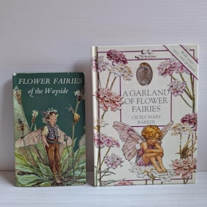 Op de afbeelding: Twee boeken met harde kaft getiteld Flower Fairies of the Wayside en A Garland of Flower Fairies door Cicely Mary Barker. Het groene boek toont een staande fee, terwijl het crèmekleurige boek een zittende fee en een kleine ingelegde hanger bevat.