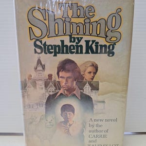 Könnte beinhalten: Ein Vintage-Taschenbuch mit dem Titel "The Shining" von Stephen King. Das Cover zeigt Illustrationen eines Mannes, einer Frau und eines Kindes, mit einem großen Haus im Hintergrund. Das Buch ist ein neuer Roman des Autors von "Carrie" und "Salem's Lot".