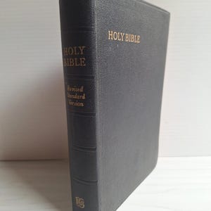 Puede incluir: Una Santa Biblia gris oscuro con letras doradas. El libro es una versión estándar revisada. El título "Holy Bible" está impreso en el lomo y la portada. Se adjunta un marcador de cinta azul.