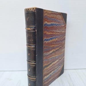 May include: Antique book with a dark leather spine and marbled cover. The spine has gold lettering and decorative bands. The cover features a swirling pattern of colors including blue, red, and brown. The book is slightly angled, showcasing its aged appearance.