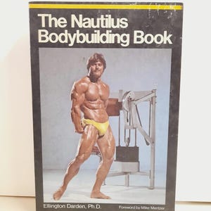 May include: The Nautilus Bodybuilding Book with a black cover. The book features a muscular man in yellow briefs posing next to a weight machine. The book's title is in white, bold font.
