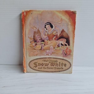 May include: A vintage hardback book titled Walt Disney's Snow White and the Seven Dwarfs. The cover features a colour illustration of Snow White surrounded by various forest animals, with a worn orange spine and a decorative oval title frame.