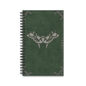 Könnte beinhalten: Spiralgebundenes Notizbuch mit dunkelgrünem Einband. Der Einband hat einen silberfarbenen Zierrahmen und ein zentrales Design einer stilisierten, symmetrischen Kreatur mit Fangzähnen. Die Seiten des Notizbuchs sind am Rücken sichtbar.