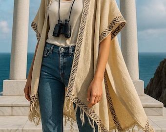 Può includere: Poncho beige con bordi a motivi geometrici e frange, indossato sopra una canotta bianca e jeans blu. Un binocolo nero è appeso al collo. Lo sfondo presenta una struttura in pietra e il mare.