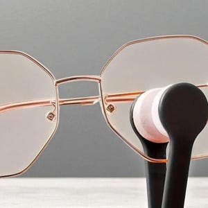 May include: Rose gold-coloured eyeglasses with a geometric frame design. The glasses are held by a black eyeglass cleaner with pink and white cleaning pads. The glasses have a modern, stylish look.