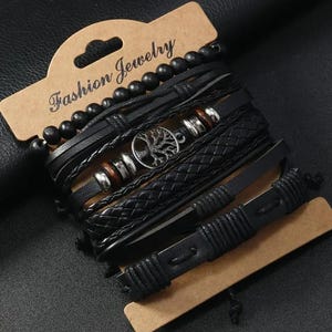 Könnte beinhalten: Ein Set schwarzer Armbänder mit verschiedenen Designs, darunter geflochtenes Leder, Perlenstränge und ein Armband mit einem Lebensbaum-Anhänger. Die Armbänder werden auf einem braunen Pappschild mit "Fashion Jewelry" präsentiert.