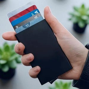 Könnte beinhalten: Ein schwarzer Kartenhalter mit mehreren Kreditkarten mit sichtbaren Logos. Der Kartenhalter ist rechteckig und scheint aus Metall zu sein. Der Hintergrund umfasst kleine Topfpflanzen.