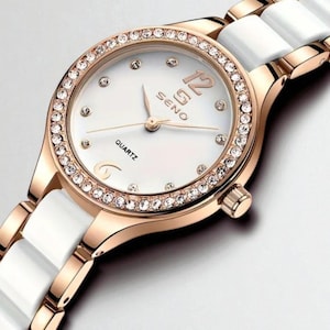 May include: A rose gold and white quartz watch with a white face and a bezel encrusted with small crystals. The watch band is white ceramic. The watch face has the word "SENO" and "QUARTZ" printed on it.