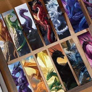 May include: A selection of dragon-themed bookmarks. Each bookmark showcases a unique dragon design in diverse colours, such as red, green, purple, and black. The dragons are illustrated in a fantasy art style, with detailed scales and wings. The bookmarks are arranged in a wooden box.