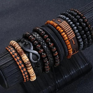 May include: A selection of bracelets in different styles. The bracelets include beaded, braided, and leather designs in shades of brown, black, and tan. Some bracelets feature wooden beads and metal accents.