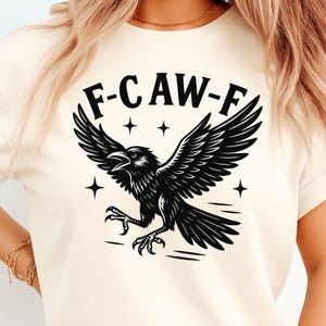 May include: A cream-coloured t-shirt with a black raven design. The raven has outstretched wings and the text "F-CAW-F" above it. Four star shapes surround the raven. The design is a graphic illustration.