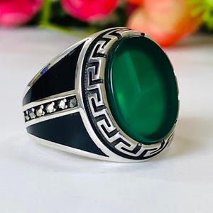 Könnte beinhalten: Silberring mit einem großen, ovalen, grünen Edelstein in der Mitte. Der Ring hat ein schwarzes Band mit kleinen, funkelnden Akzenten und einem dekorativen Rand um den grünen Stein.