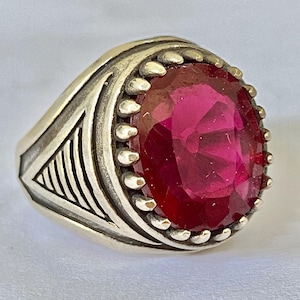 May include: A silver ring featuring a large, oval-cut, deep pink gemstone set in a pronged bezel. The band displays a textured, geometric pattern with engraved lines along the sides, creating a structured appearance around the central stone.