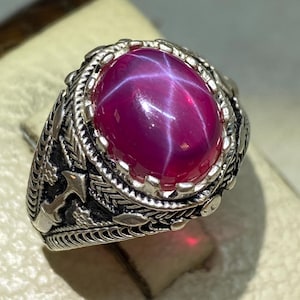Puede incluir: Anillo de plata con una gran gema de rubí estrella ovalada. El rubí es de color magenta intenso con un patrón de estrella de seis rayos. El anillo tiene un diseño ornamentado con motivos florales y de hojas.