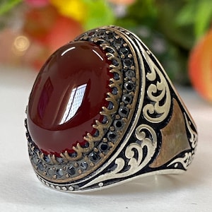 Puede incluir: Anillo de plata con una piedra preciosa ovalada de color rojo intenso. La piedra está rodeada por un borde decorativo de pequeñas piedras negras y un detallado diseño de filigrana plateada. El anillo tiene una estética vintage.