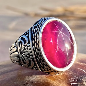 Könnte beinhalten: Ein silberner Ring mit einem ovalen, tiefroten Edelstein. Der Edelstein weist ein sternförmiges Muster auf. Das Band des Rings ist mit detaillierten Schnitzereien verziert. Der Ring ist vor einem unscharfen Hintergrund platziert.