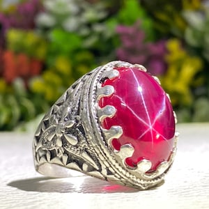 May include: A silver ring featuring a large, oval-shaped, deep red star ruby cabochon with a visible six-rayed star effect. The metal band displays intricate, raised floral and geometric carvings along the sides and a scalloped bezel setting.