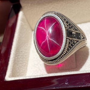 May include: A silver ring featuring a large, oval, cabochon-cut magenta stone with a visible six-rayed star effect. The metal band displays intricate, textured, and oxidized detailing along the sides, framing the central gemstone securely.