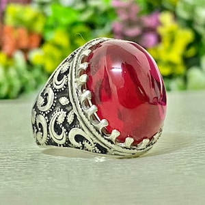 Pode incluir: Um anel de prata com uma grande pedra vermelha translúcida de corte cabochão oval, fixada numa cravação de garras. A banda tem um acabamento em prata oxidada e apresenta padrões intrincados de volutas em relevo nas laterais.