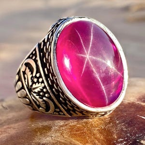 May include: A silver ring with an oval, vibrant pink gemstone. The gemstone displays a six-rayed star pattern. The ring's band features intricate, detailed carvings, adding to its ornate design. The ring is set against a blurred background.