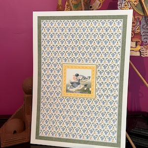 Op de afbeelding: Een ingelijste prent met een eendenillustratie in een gouden lijst, tegen een achtergrond van blauwe en gouden bloempatronen. De prent is omlijst door een groene lijst, waardoor een decoratief kunstwerk ontstaat.